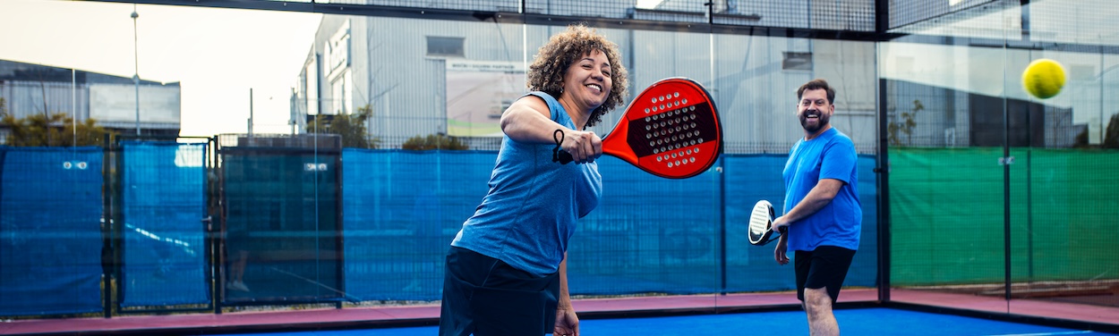Woman Playing Pickleball with Partner A smiling woman with curly hair in a blue shirt is in mid-swing, playing padel with a red pickleball racquet on a blue court. A man in a blue shirt and black shorts stands behind her, also smiling, holding his pickleball racquet. A bright yellow padel ball is visible in the air.